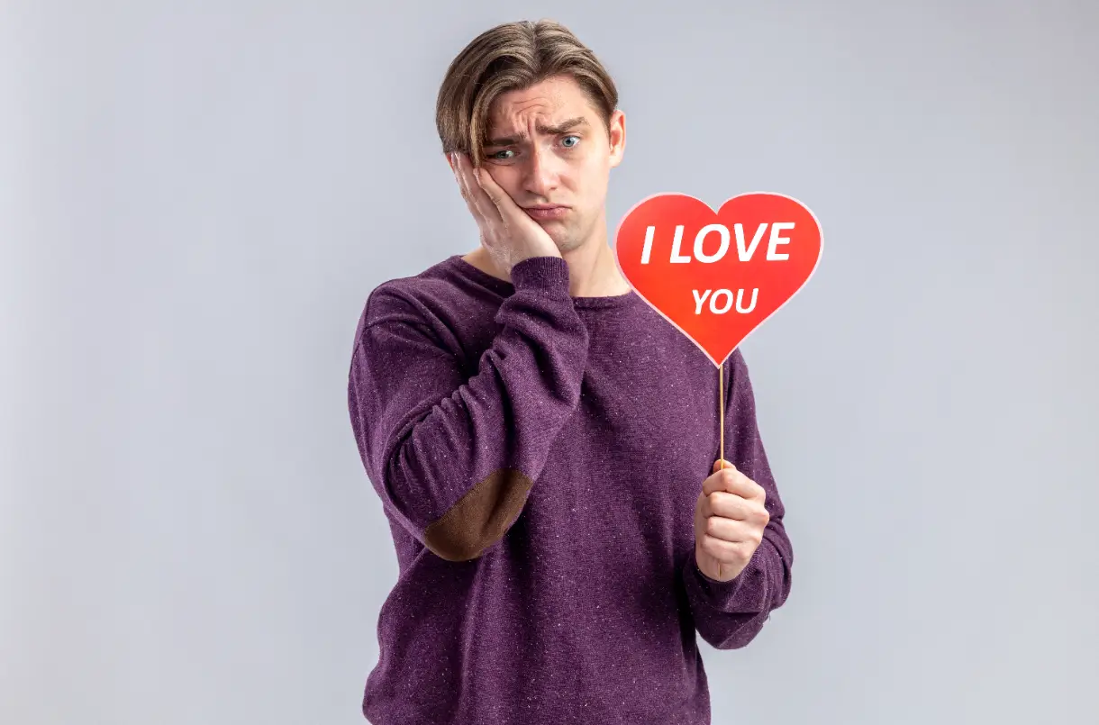 Elinde “I love you” yazılı kalp tabelası tutan, eli yanağında mutsuz ve düşünceli genç adam, sevgi ile obsesif aşk arasındaki fark